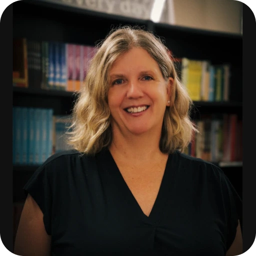 A smiling woman with blonde, wavy hair and a black top poses in front of a bookshelf filled with books.