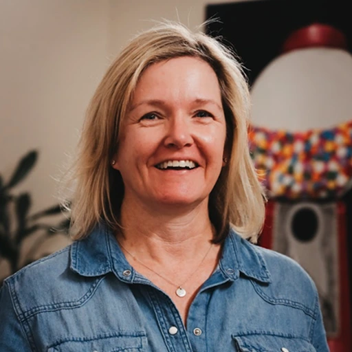 A smiling woman with blonde hair wearing a denim button-up shirt, with a gum ball fundraiser tracker in the background.