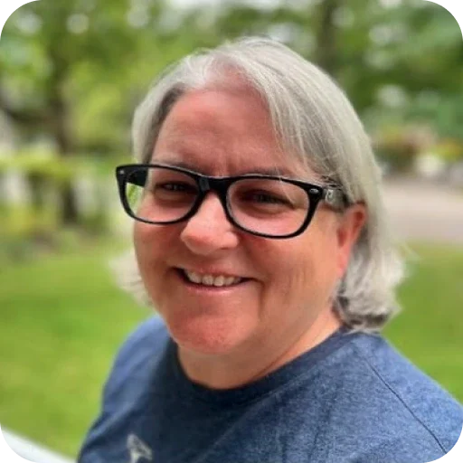 A smiling woman with glasses and short grey hair wearing a blue t-shirt outdoors, with green trees blurred in the background.