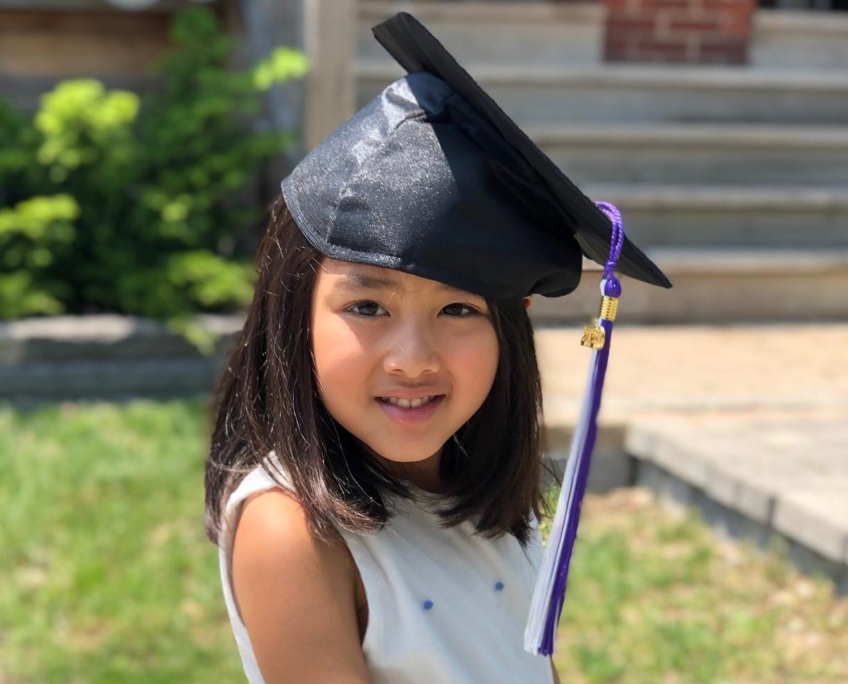A smiling young girl wears a black graduation cap with a purple and white tassel outdoors.