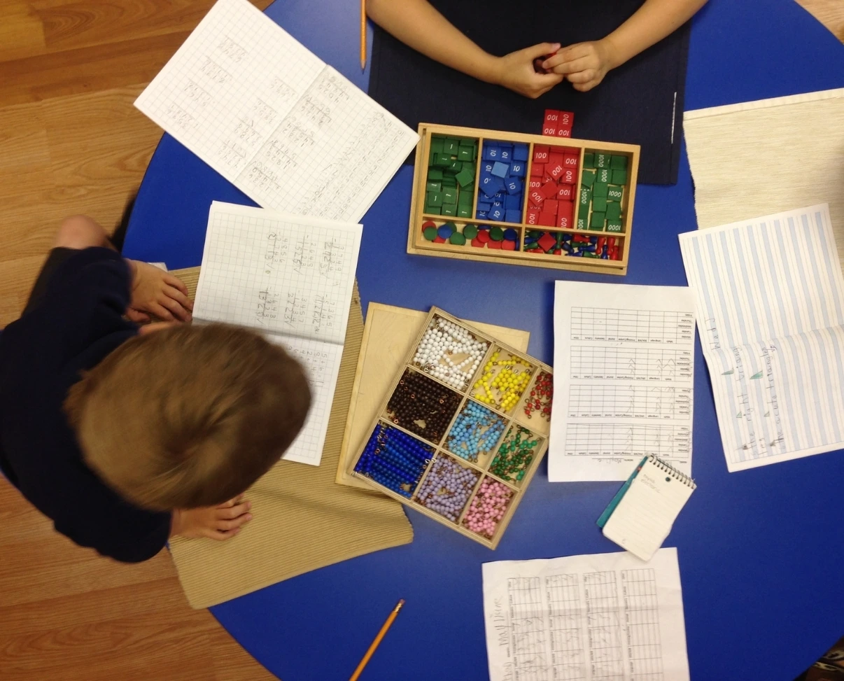 An overhead view of students at a blue table working on math assignments with notebooks, coloured beads, and counting tiles.
