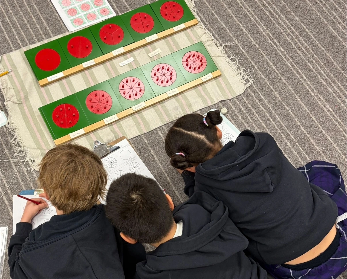 Three students sit on the floor working on a fraction lesson with worksheets and wooden circle fraction inserts.
