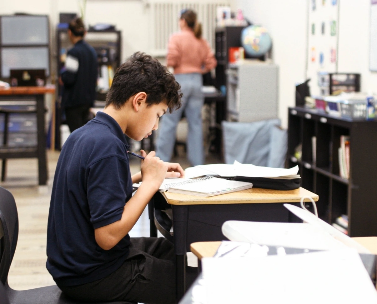 A student in a blue polo shirt sits at his desk, focused on writing in a notebook in a busy classroom.