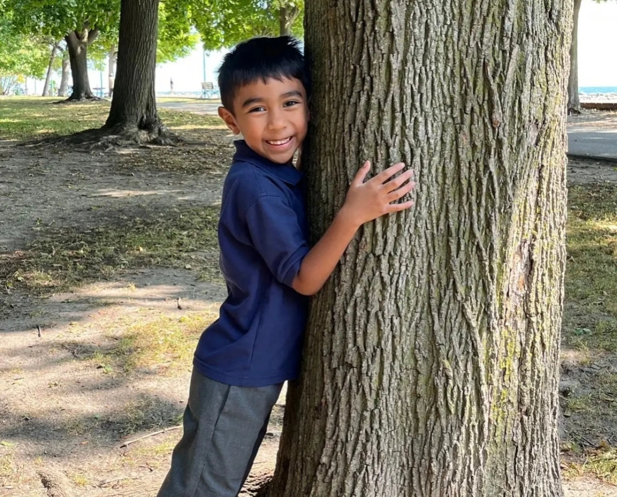 A smiling young boy in a blue polo uniform hugs a large tree trunk in a grassy park setting.