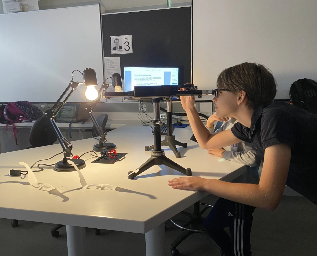 A student with glasses leans forward to look through a lab spectrometer aimed at a glowing lightbulb on a desk.