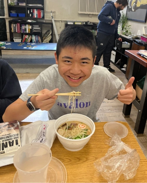 A smiling student in an Avalon t-shirt eats ramen with chopsticks and gives a thumbs-up at a classroom table.