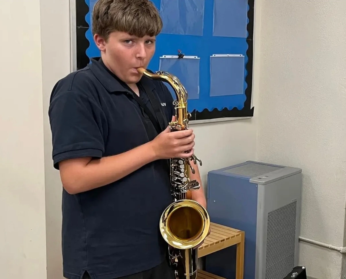 A student in a dark polo shirt stands indoors playing a saxophone, looking toward the camera.