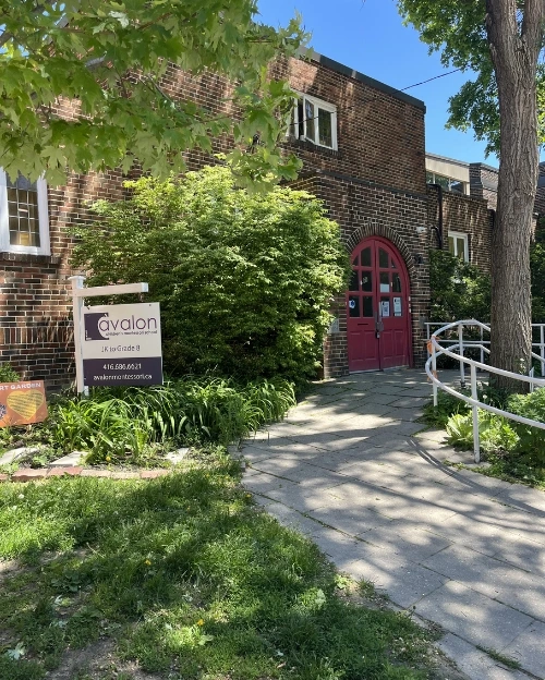 A stone path leads to the arched red doors of a brick Avalon school building, past a sign on the grassy lawn.