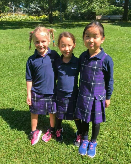 Three girls smile on a sunny lawn wearing matching purple and navy plaid school uniforms.