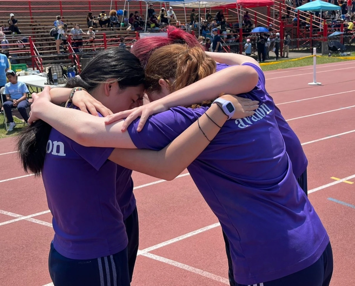 Three students wearing purple "avalon" shirts huddle together arm-in-arm on a running track during a sports event.