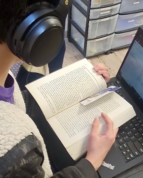 A student wearing large headphones holds an open book over a laptop keyboard while reading.