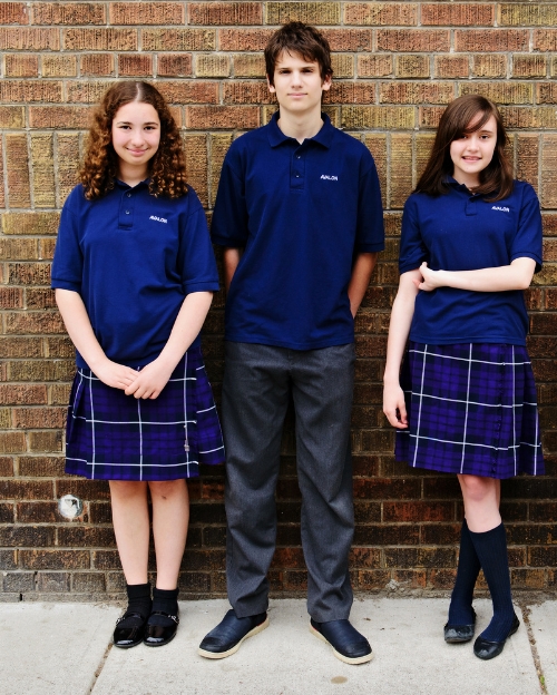 Three students pose in front of a brick wall wearing navy Avalon polo shirts with plaid skirts or grey trousers.
