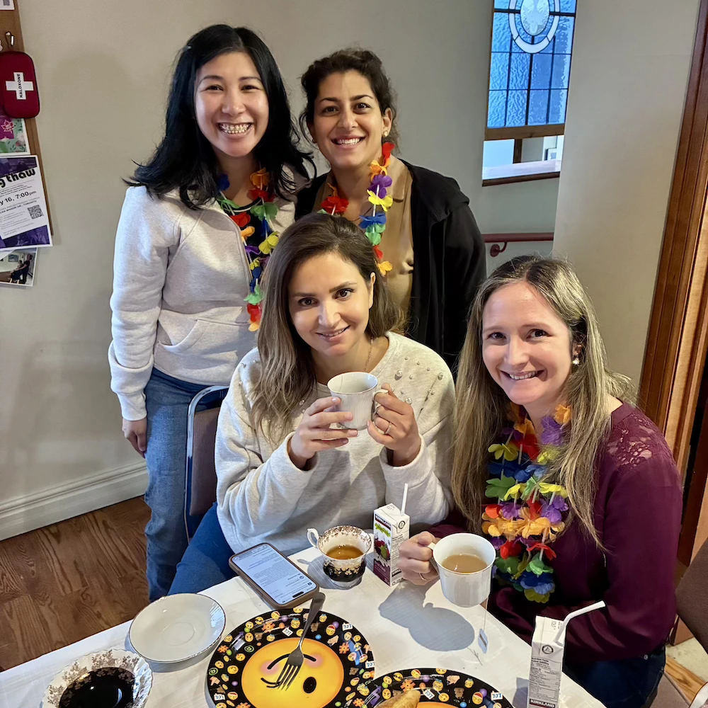 Four smiling women gather around a table with tea cups and juice, some wearing colorful flower leis.